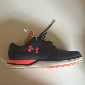 Golfskor från Under Armour - Snygga mörkblåa golfskor från Under Armour med rosa detaljer och snörning. Perfekta för träning med en bekväm sula och sportig design. Känns som en storlek 39