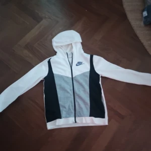 Grå vit Nike hoodie - Säljer en snygg vit och grå hoodie från Nike med svarta detaljer. Säljer den för den är för liten för mig och den är i bra skick