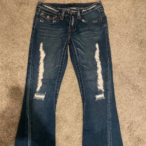 Blå bootcut jeans från True Religion - MÅTT: längd från midja: 99cm. inerbens mått: 78cm. midja: 72cm.                                              Snygga blå bootcut jeans från True Religion med slitna detaljer på framsidan. Jeansen har klassiska femfickor och märkets ikoniska broderi på bakfickorna. 