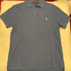 Blå pikétröja från Ralph Lauren - Sparsamt använd. Kameran gör att den blåa färgen ser sämre ut, är mer fyllig i färgen. Snygg blå pikétröja från Ralph Lauren i custom slim fit. Tröjan har en klassisk krage med två knappar och det ikoniska broderade logotypen på bröstet. Perfekt för en stilren look!