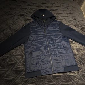 J.LINDEBERG marinblå hybridjacka - Snygg mörkblå hybridjacka huva från J.LINDEBERG. Jackan har en quiltad design på framsidan och dragkedja. Köpt i december 2023 och därmed i väldigt bra skick. Passar som M. NYPRIS: 2499kr