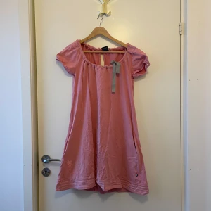 Rosa klänning från House of Lola - Charmig rosa klänning från House of Lola med korta ärmar och rund halsringning. Klänningen har fickor.