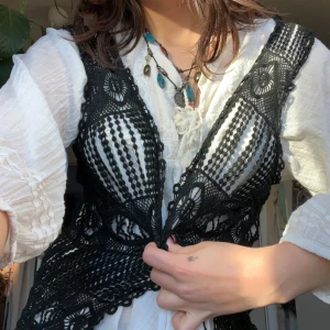 Svart spetsväst - Snygg svart spetsväst med intrikat mönster. Perfekt för att ge en bohemisk touch till din outfit. Västen är öppen framtill och har ett vackert, detaljerat spetsmönster. Det finns ingen knapp men det är lätt att lägga till.