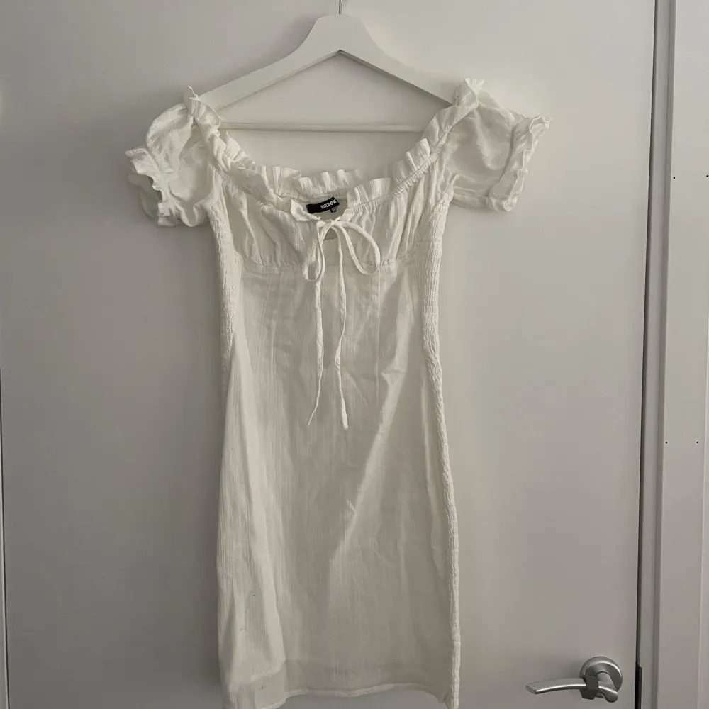 En aldrig använd söt vit klänning från BikBok i storlek XS. Klänningen har en offshoulder-design med volangdetaljer och knyt framtill. Perfekt för sommaren, skolavslutning eller student!. Mekot.