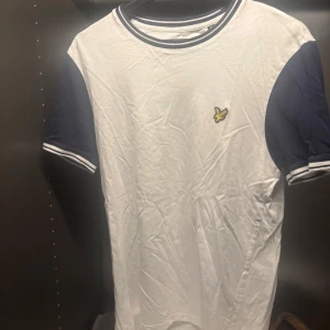 Vit och marinblå t-shirt från Lyle & Scott - Säljer en stilren vit t-shirt från Lyle & Scott med marinblå ärmar och krage. T-shirten har en liten broderad logga på bröstet och är kortärmad. Perfekt för en avslappnad look.