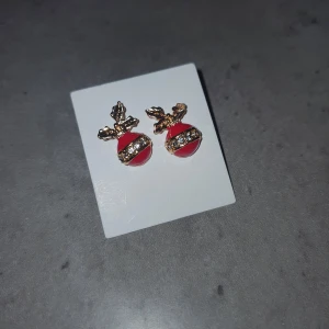 Örhängen med renmotiv - Charmiga örhängen i rött och guld med ett renmotiv. De har en glittrig detalj i mitten som ger en festlig känsla. Perfekta för att lysa upp din outfit under julen.