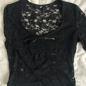 Svart spetsblus med rosett - Elegant svart spetsblus med v-ringning och en söt rosett framtill. Blusen har långa ärmar och ett genombrutet mönster som ger en romantisk känsla. Perfekt för att lägga till en touch av elegans till din outfit.