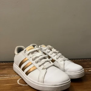 Vita och guldiga sneakers från Adidas - Snygga vita sneakers från Adidas med guldiga detaljer. Skorna har de klassiska tre ränderna i guld på sidan och snörning framtill. Perfekta för en stilren look. De är använda en enda gång och är 10/10 i skick inga defekter eller fläckar alls. Om du undrar något fråga 