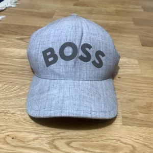 Grå keps från Boss - Snygg grå keps från Boss med texten 'BOSS' i svart på framsidan. Kepsen har en klassisk design med böjd skärm och justerbar passform. Perfekt för en stilren look.nypris 500kr!