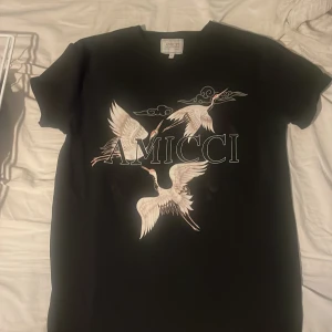 Svart t-shirt med tryck från Amicci - Snygg svart t-shirt från Amicci med ett coolt tryck av fåglar och texten 'AMICCI' på framsidan. Perfekt för en stilren look.