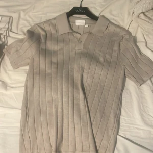 Beige pikétröja med ribbad design - Snygg beige pikétröja med ribbad design och korta ärmar. Perfekt för en stilren och avslappnad look. Tröjan har en klassisk krage och knappar framtill.