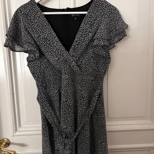 Svart och vit blommig klänning från Gina Tricot - Säljer en söt svart och vit blommig klänning från Ghica London. Klänningen har en v-ringad design med kortärmade volanger och ett knytband i midjan. Perfekt för en somrig look! 🌸