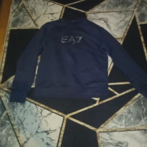 Mörkblå tröja från Emporio Armani - Säljer en stilren mörkblå tröja från Emporio Armani med EA7-logga på bröstet. Tröjan har långa ärmar och en klassisk passform. Perfekt för en avslappnad och trendig look.