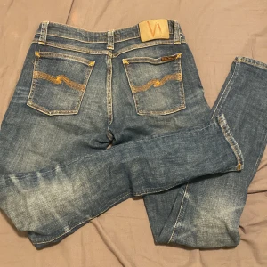Blå jeans från Nudie Jeans - Säljer ett par snygga blå jeans från Nudie Jeans med klassisk femficksdesign och orange sömmar. Jeansen har en normal passform och är tillverkade i slitstarkt denim. Perfekta för en avslappnad stil.