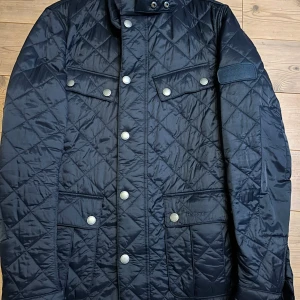 Svart quiltad jacka från Barbour - Säljer en stilren svart quiltad jacka från Barbour International. Jackan har långa ärmar och knappar framtill samt på fickorna. Perfekt för kyligare dagar med sin klassiska design och praktiska detaljer.