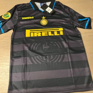 Svart och grå Inter fotbollströja från Umbro - Säljer en svart och grå randig Inter fotbollströja från Umbro med gula detaljer. Tröjan har korta ärmar och Pirelli som sponsorlogga på framsidan. Nummer 10 och namn på baksidan. Perfekt för samlare eller fans!