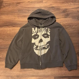 Grå hoodie med Misfits-tryck  - Säljer en grå hoodie med ett stort Misfits-tryck på framsidan. Hoodien har dragkedja och en klassisk huva.