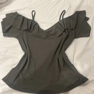 Grå topp med volangdetaljer - Snygg grå topp med smala axelband och volangdetaljer längs halsringningen. Perfekt för en stilren look med en touch av elegans.