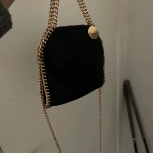 Svart axelväska med kedjedetaljer - Snygg svart axelväska med guldfärgade kedjedetaljer och en rund guldfärgad knapp. Perfekt för att ge en stilren touch till din outfit. Väskan har en elegant design och passar bra för olika tillfällen.finns en liten defekt (skriv för bild)