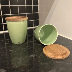 Gröna muggar med trälock från Kosta Boda - Säljer två stilrena gröna muggar från Kosta Boda med matchande trälock. Perfekta för att hålla drycken varm eller förvara småsaker. Muggarna har en elegant form och är tillverkade i keramik med ett naturligt trälock.
