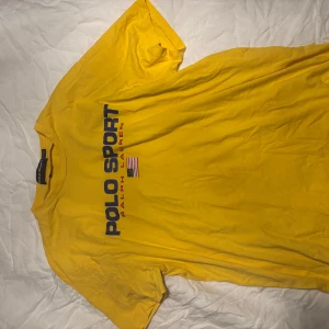 Gul t-shirt från Polo Sport - Säljer en gul t-shirt från Polo Sport Ralph Lauren med blå och röd logga på framsidan. T-shirten har en klassisk passform och är kortärmad. Perfekt för en sportig look!