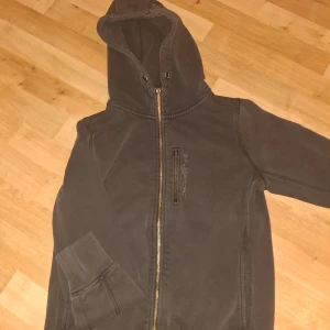 Svart huvtröja från Sail Racing - Säljer en svart huvtröja från Sail Racing med dragkedja och broderad logga på bröstet. Tröjan har en justerbar huva och långa ärmar med ribbade muddar. Perfekt för en avslappnad stil.