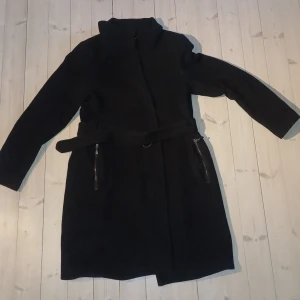 Svart kappa från Esprit - Snygg svart kappa från Esprit med dragkedja och bälte i midjan. Perfekt för kyligare dagar med sin stilrena design och långa ärmar. Kappan har praktiska fickor med dragkedjor.