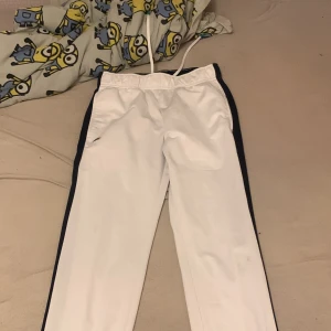 Vita sportbyxor från Lacoste - Snygga vita sportbyxor från Lacoste med svarta sidopaneler och gröna detaljer baktill. Byxorna har en elastisk midja och det klassiska Lacoste-logotypen på framsidan. Perfekta för en sportig look. Också från Frankrike 🇫🇷 