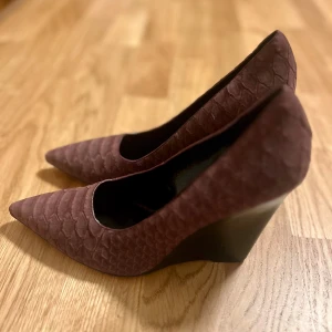 Vinröd pumps Burberry med ormskinnsmönster - Snygga vinröd pumps med spetsig tå och ormskinnsmönster. Perfekta för att ge en elegant touch till din outfit. Klackarna är stabila och ger bra höjd.