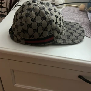 Mönstrad keps från Gucci - Snygg keps från Gucci med det ikoniska GG-mönstret i svart och beige. Kepsen har en justerbar rem baktill och är perfekt för att ge en lyxig touch till din outfit. Kvitto och boxen igår o allt som tillhör kepsen