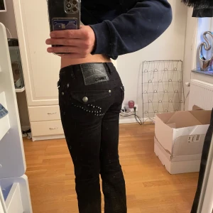 Svarta jeans med nitar - Snygga svarta jeans med tight passform och coola nitar på bakfickorna. De har en lågmidjad design och är perfekta för en trendig look. Jeansen har knappar och dragkedja framtill.