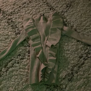 Beige ribbad tröja med volangdetaljer - Säljer en snygg beige ribbad tröja med volangdetaljer längs framsidan. Tröjan har långa ärmar och en tight passform. Passar bra till både jeans och kjol. Hör av dig vid intresse!