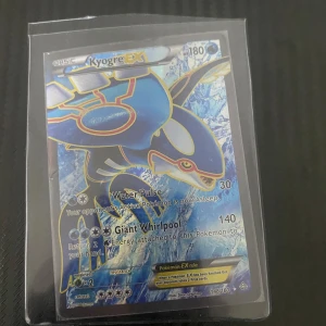 Kyogre EX Pokémonkort - Säljer ett Kyogre EX Pokémonkort med 180 HP. Kortet har attacker som Water Pulse och Giant Whirlpool. Det är en del av en samling och har ett holografiskt utseende. Kortet är skyddat i en plastficka.