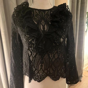 Svart spetsblus med volangdetaljer - Elegant svart spetsblus med vackra volangdetaljer framtill. Blusen har långärmade ärmar och ett intrikat spetsmönster som ger en sofistikerad look. Perfekt för att lyfta vilken outfit som helst.