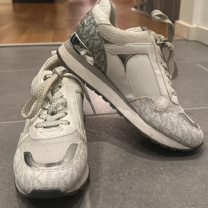 Vita sneakers från Michael Kors - Snygga vita sneakers från Michael Kors med silverdetaljer och snörning. Skorna har en kombination av skinn och syntetmaterial med en mönstrad sula. Perfekta för en stilren look. Använt 2 gånger, inget fel på dem. 