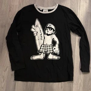 Svart långärmad tröja med surfmotiv från bpc - Cool svart långärmad tröja från bpc med ett stort tryck av en surfare på framsidan. Perfekt för en avslappnad stil med en touch av surfkultur. Tröjan har en rund halsringning med kontrasterande vit kant.