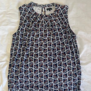 Mönstrad ärmlös topp från Stockh lm - Snygg ärmlös topp från Stockh LM med ett unikt mönster i blått, vitt och rött. Perfekt för varma dagar eller som ett lager under en kavaj. Toppen har en rund halsringning och en knappdetalj i nacken.