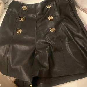 Svart skinnkjol med guldknappar - Snygg svart skinnkjol med sex dekorativa guldknappar framtill. Kjolen har en stilren design och är perfekt för att ge en edgy touch till din outfit. Den är kort och passar bra till olika tillfällen. Ett måste för garderoben!