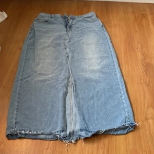 Lång jeanskjol med slitna detaljer - Säljer en cool lång jeanskjol i ljusblå denim med slitna kanter. Kjolen har en klassisk femficksdesign och en rak passform. Perfekt för en avslappnad stil.