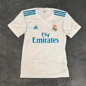 Vit Real Madrid fotbollströja från Adidas - Säljer en vit Real Madrid fotbollströja från Adidas med korta ärmar och blå detaljer. Tröjan är knappt använd och köptes 2017. Priset kan diskuteras 