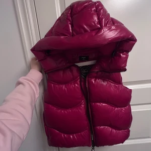 Röd dunväst från Superdry - Säljer en snygg röd dunväst från Superdry med huva och dragkedja. Perfekt för kyliga dagar när du vill hålla värmen utan att kompromissa med stilen. Västen har en cool XP-D logga på bröstet.