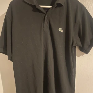 Svart pikétröja från Lacoste - Säljer en klassisk svart pikétröja från Lacoste med den ikoniska krokodilloggan på bröstet. Tröjan har korta ärmar och en stilren krage. Perfekt för en avslappnad men ändå stilfull look. Den är i storlek 4 så tror de motsvarar runt m/l