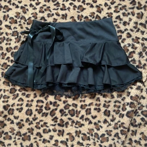 Svart volangkjol med spetsdetaljer - Snygg svart kortkjol med volanger och spetsdetaljer. Kjolen har en dekorativ rosett i midjan som ger en extra touch. Perfekt för att skapa en chic look.