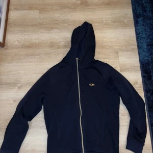 Svart hoodie från boss - Snygg svart hoodie från boss med dragkedja framtill. Tröjan har långa ärmar och en praktisk huva. Perfekt för en avslappnad stil.