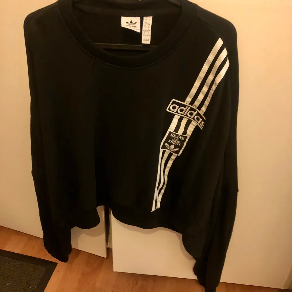 Snygg svart tröja från Adidas med de klassiska tre ränderna i vitt på framsidan. Tröjan har en rund halsringning och långa ärmar. Perfekt för en sportig look eller avslappnad stil.. Neuletakit & Villapaidat.