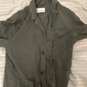 Grå overshirt från A Day's March - Säljer en mörk grön overshirt från A Day's March. Den har en knappstängning framtill och en bröstficka med knapp. Perfekt för lager-på-lager-stil. Passar bra till både jeans och chinos.