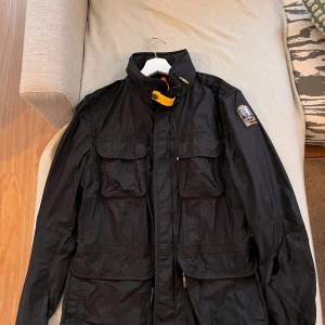 Svart jacka från Parajumpers - Säljer en svart jacka från Parajumpers med flera fickor och en cool patch på ärmen. Jackan har en hög krage med en orange detalj och dragkedja framtill. Perfekt för höst och vår!