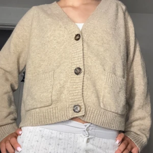 Beige kofta med knappar - Mysig beige kofta med stora knappar framtill. Den har långa ärmar och ribbade detaljer vid ärmslut och nederkant. Perfekt för kyligare dagar.