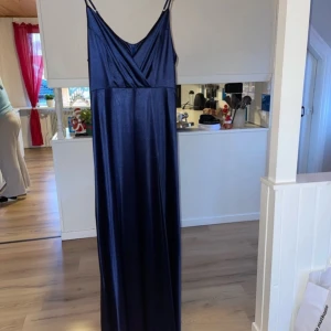 Mörkblå långklänning från Muxxn - Elegant mörkblå långklänning från Muxxn i ett glansigt material. Klänningen har smala axelband och en v-ringad design som ger en stilren look. Använd vid bara ett tillfälle så fortfarande i ett bra skick
