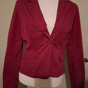 Vinröd kavaj från Top Secret - Snygg vinröd kavaj från Top Secret i storlek 38. Kavajen har en klassisk design med en knapp framtill och långa ärmar med tre knappar vid ärmsluten. Perfekt för en stilren look.